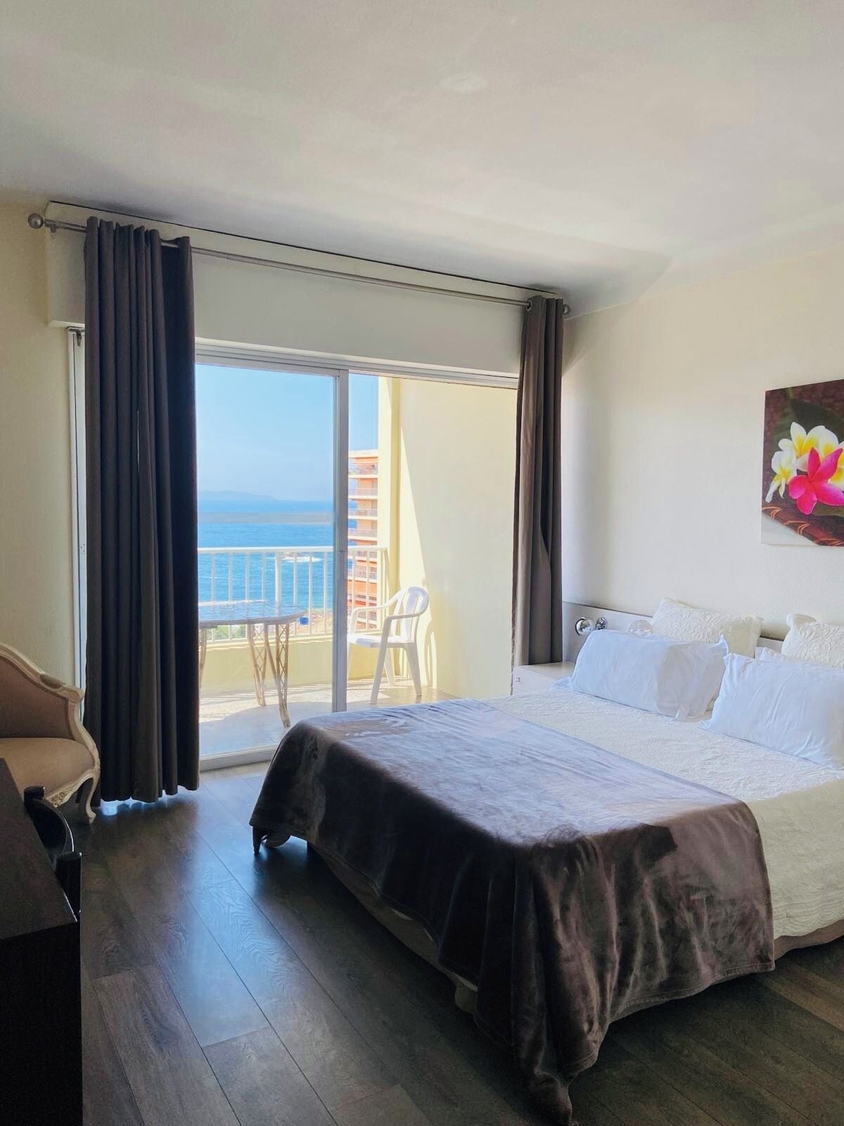 Una camera d'albergo con un letto, una sedia, un balcone vista mare e un quadro appeso al muro.