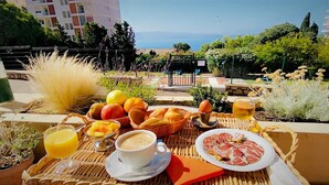 Un plateau de petit-déjeuner comprenant des fruits, des viennoiseries, du café et une assiette de charcuteries, le tout disposé sur un balcon offrant une vue sur la ville et la mer.