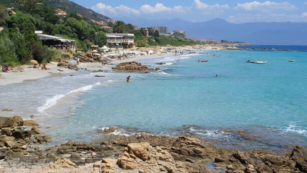 Une plage côtière aux rivages rocheux, avec une eau d’un bleu limpide et des bâtiments qui bordent la côte.