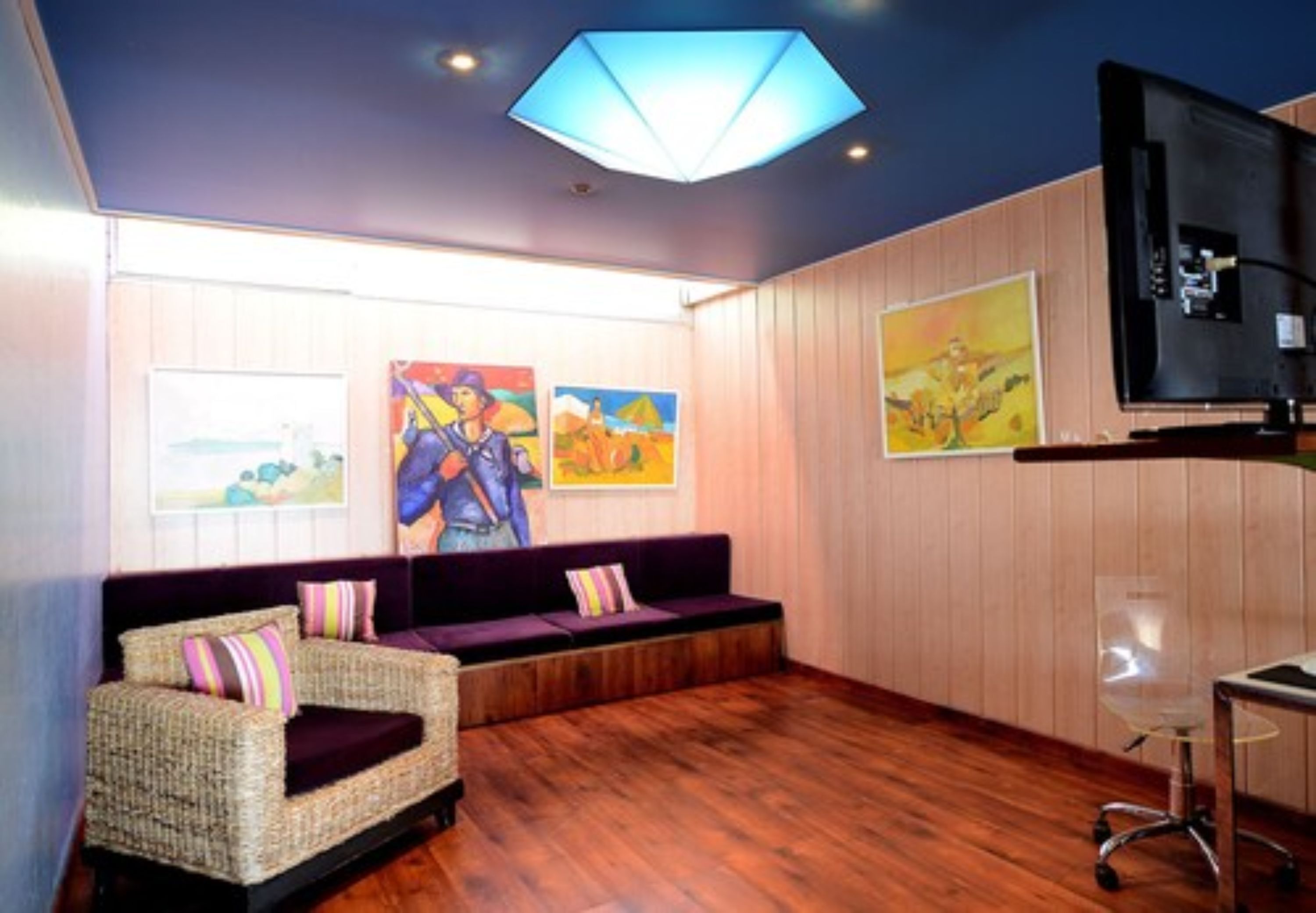 A room with a wooden floor, a wicker sofa with cushions, a television, and artwork on the walls.