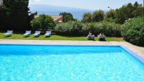 Une piscine d’un bleu éclatant, dotée de chaises longues et d’une table aménagée pour les repas, le tout entouré de verdure et offrant une vue sur la mer.