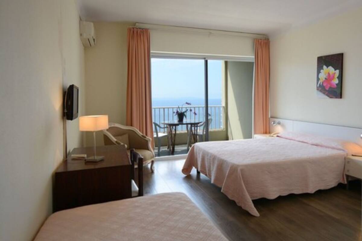 A hotel room with two beds, a desk, a chair, a balcony with a table and chairs, and a painting on the wall.