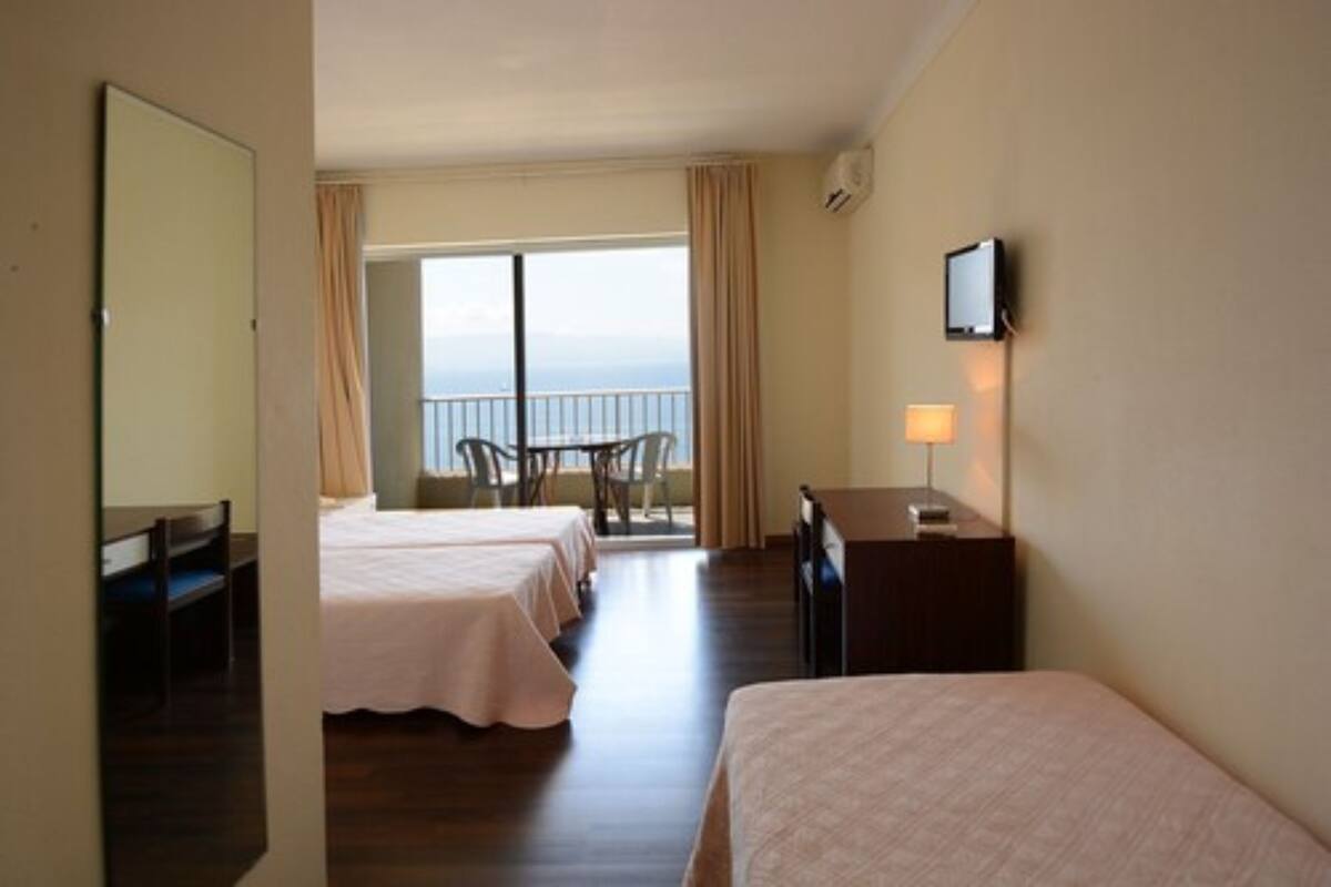 A hotel room with two beds, a desk, a chair, a TV, and a balcony with a view.