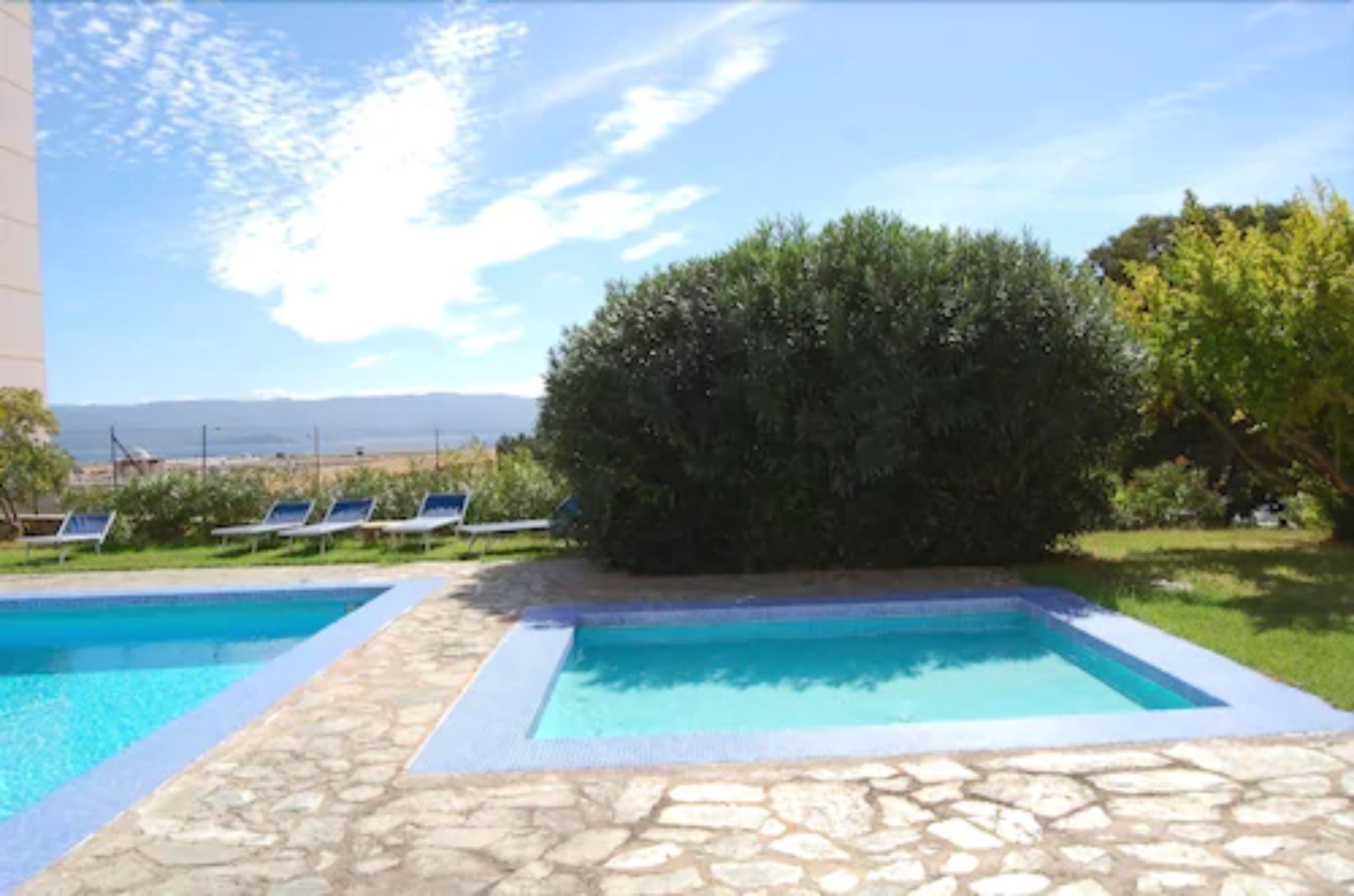 A swimming pool area with two pools, stone flooring, and a view of the landscape.