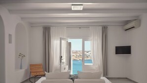 A hotel room with a bed, a TV, a wooden chair, and a view of the sea. - Pietra e Mare - Mykonos Moments by Mr and Mrs White (Mykonos)