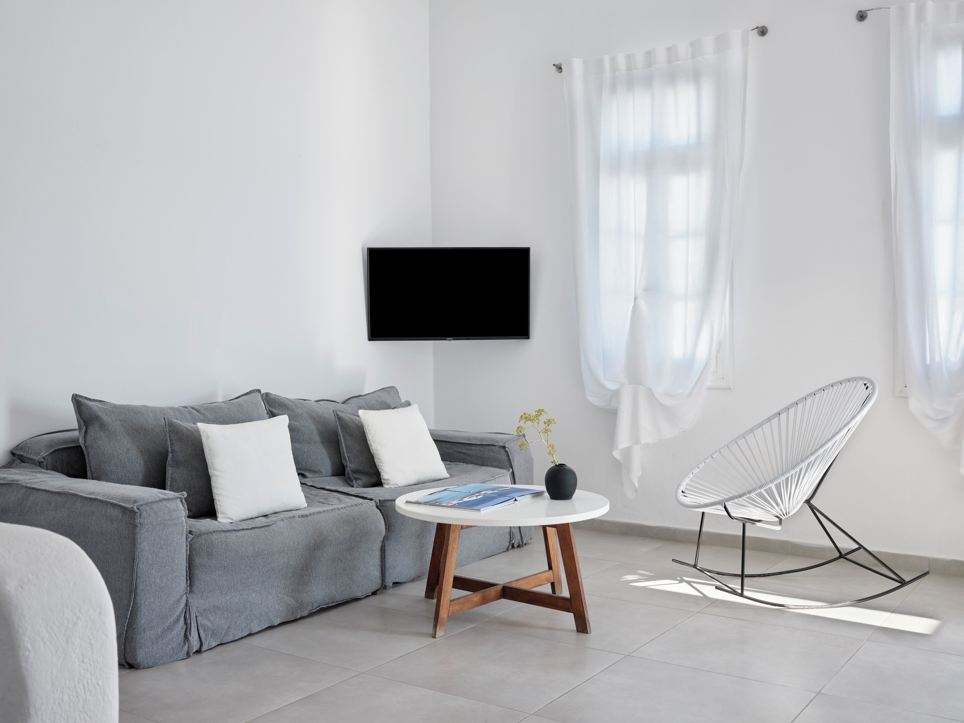 En moderne stue med en grå sofa, et rundt træbord, en hvid stol og et fladskærms-tv på væggen.