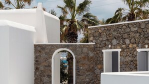 A white building with a stone wall, an arched doorway, and palm trees in the background. - Pietra e Mare - Mykonos Moments by Mr and Mrs White (Mykonos)