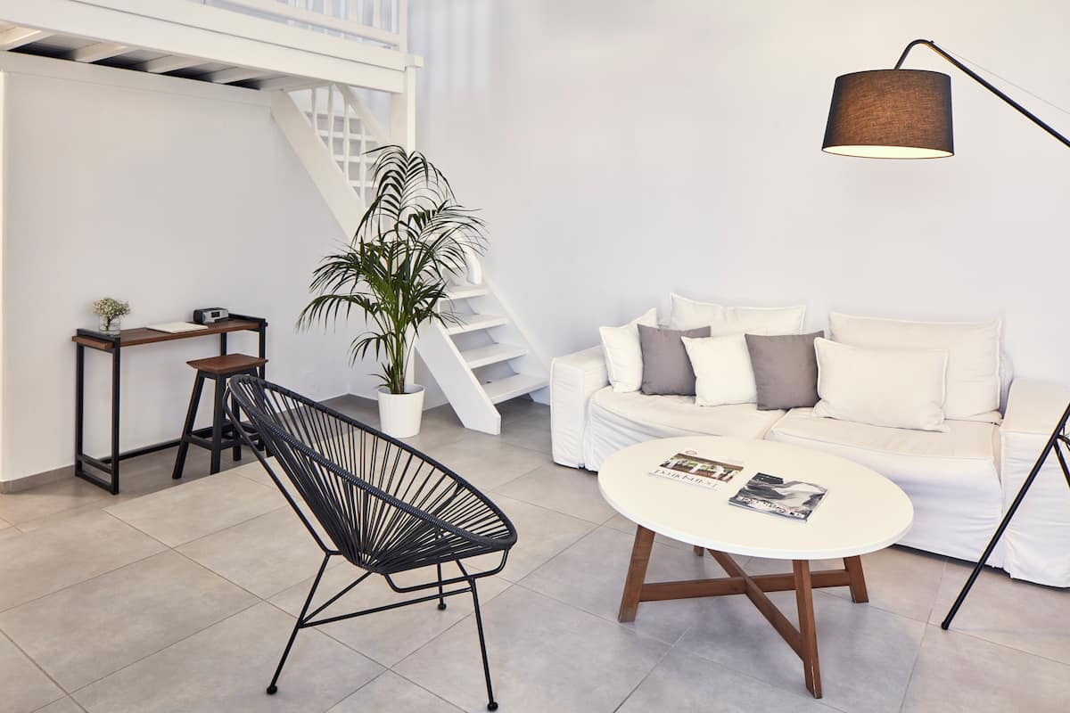 Una sala moderna con un sofá blanco, un sillón negro, una mesa de centro redonda, una lámpara de pie, una escalera y una planta en maceta.