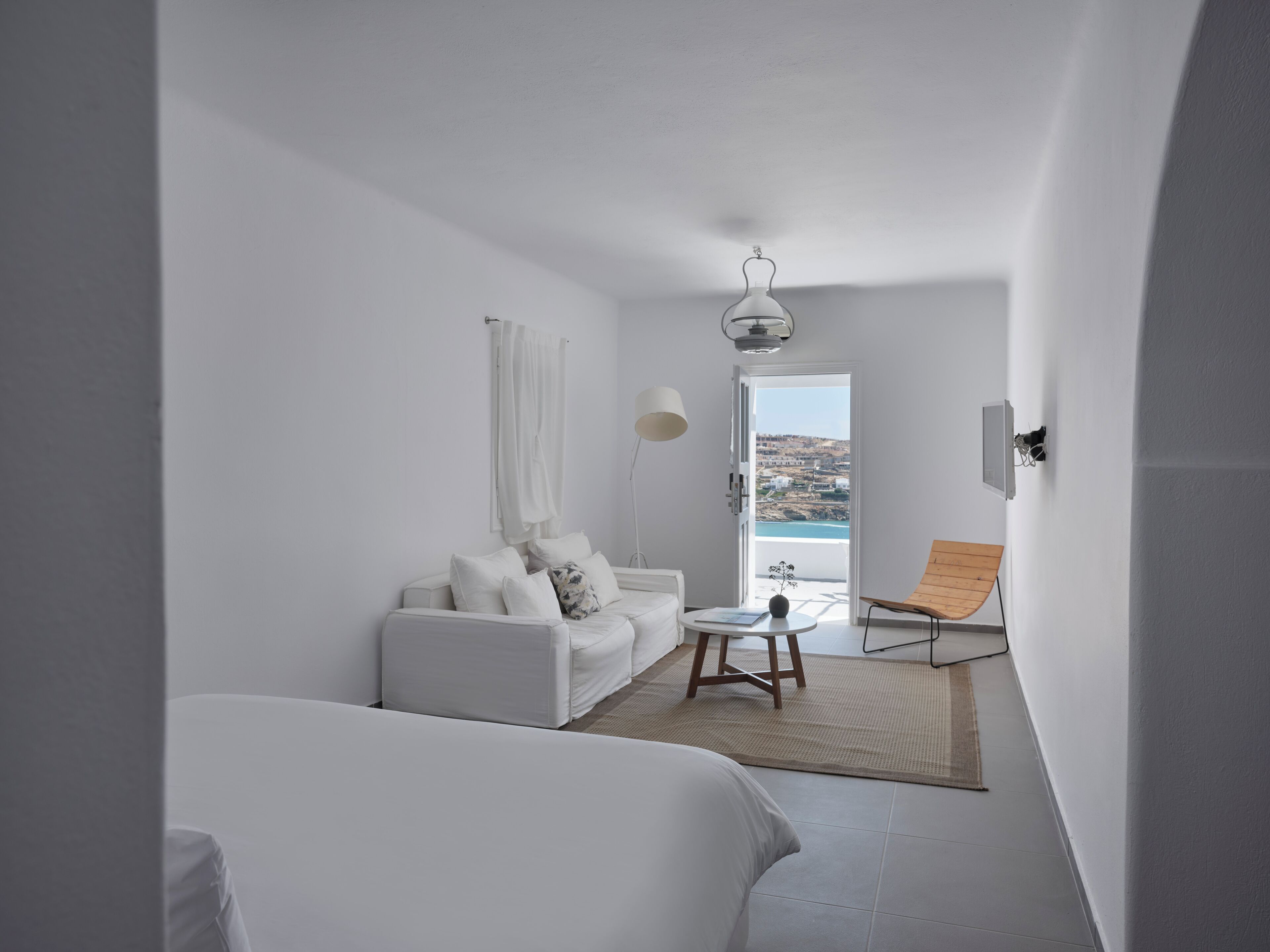 Une pièce moderne et minimaliste, avec un canapé blanc, une petite table et une chaise. Une grande fenêtre offre une vue sur la mer et les collines.