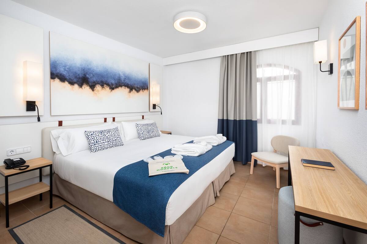 Une chambre d’hôtel avec un lit bien fait, un bureau, une chaise et une grande fenêtre avec des rideaux.