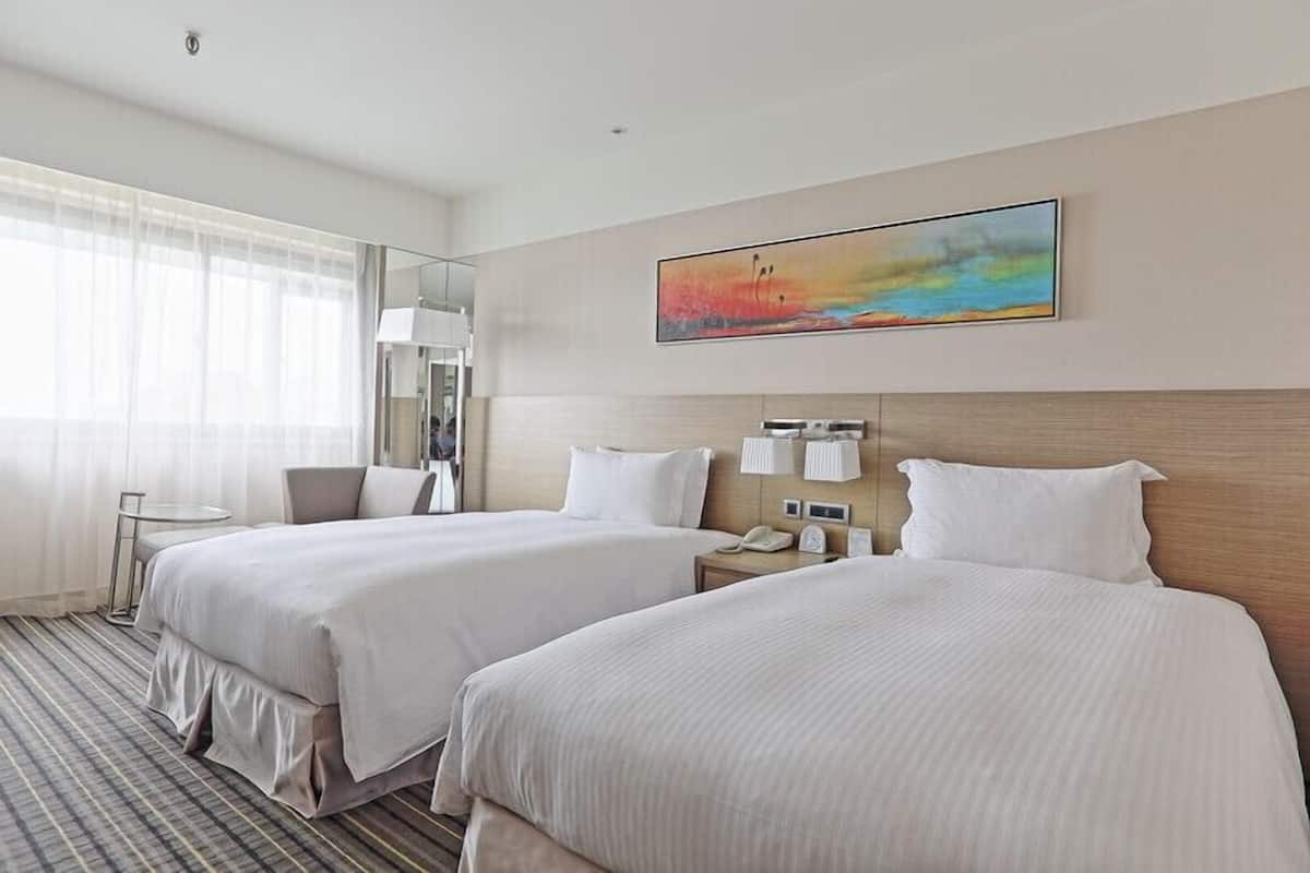A hotel room with two beds, a bedside table with a lamp, a painting on the wall, and a window with sheer curtains.