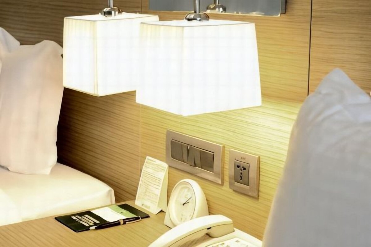 Habitación de hotel con cama, mesita de noche con un libro, teléfono, reloj y panel de control.
