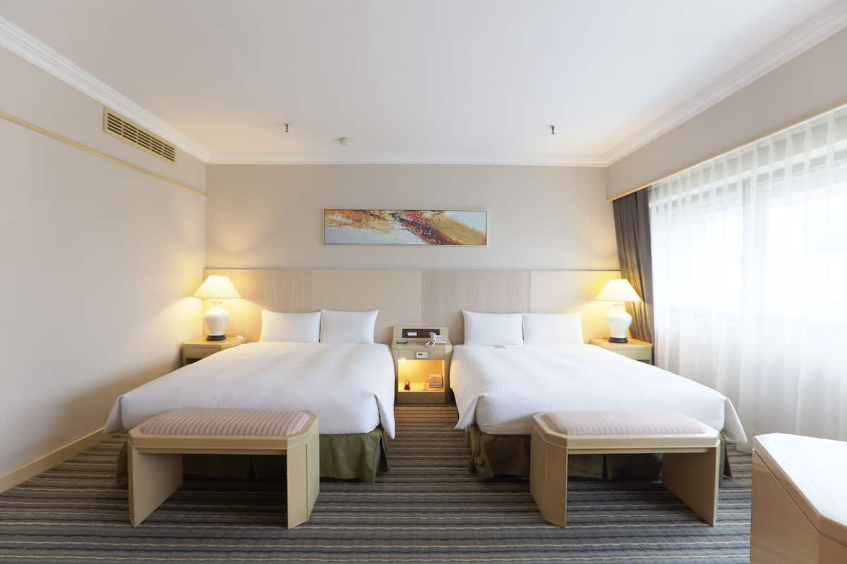 A hotel room with two beds, a desk, a chair, a lamp, a painting on the wall, and a window with curtains.