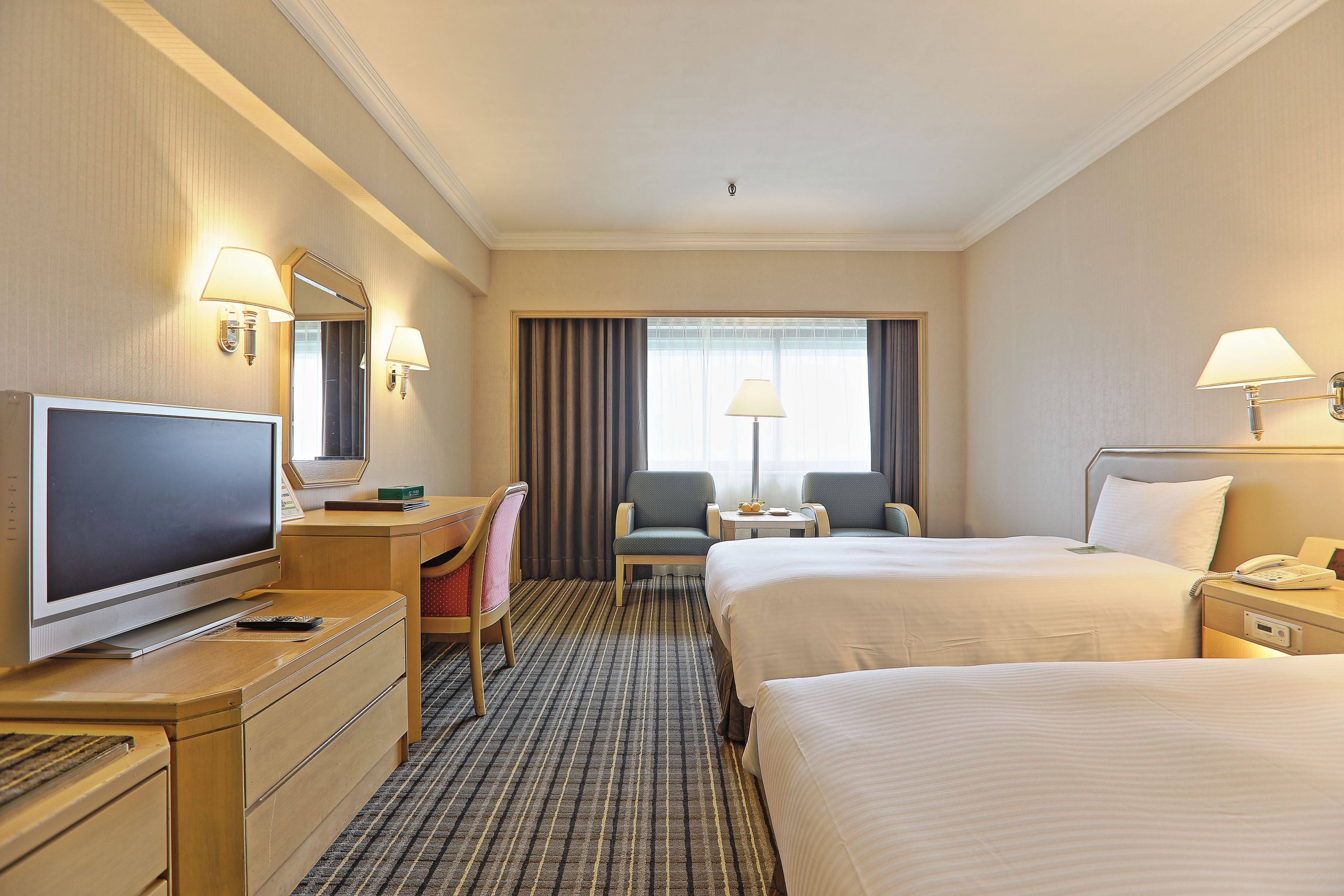 A hotel room with a TV, two beds, a desk, a chair, a small table, and a window with curtains.