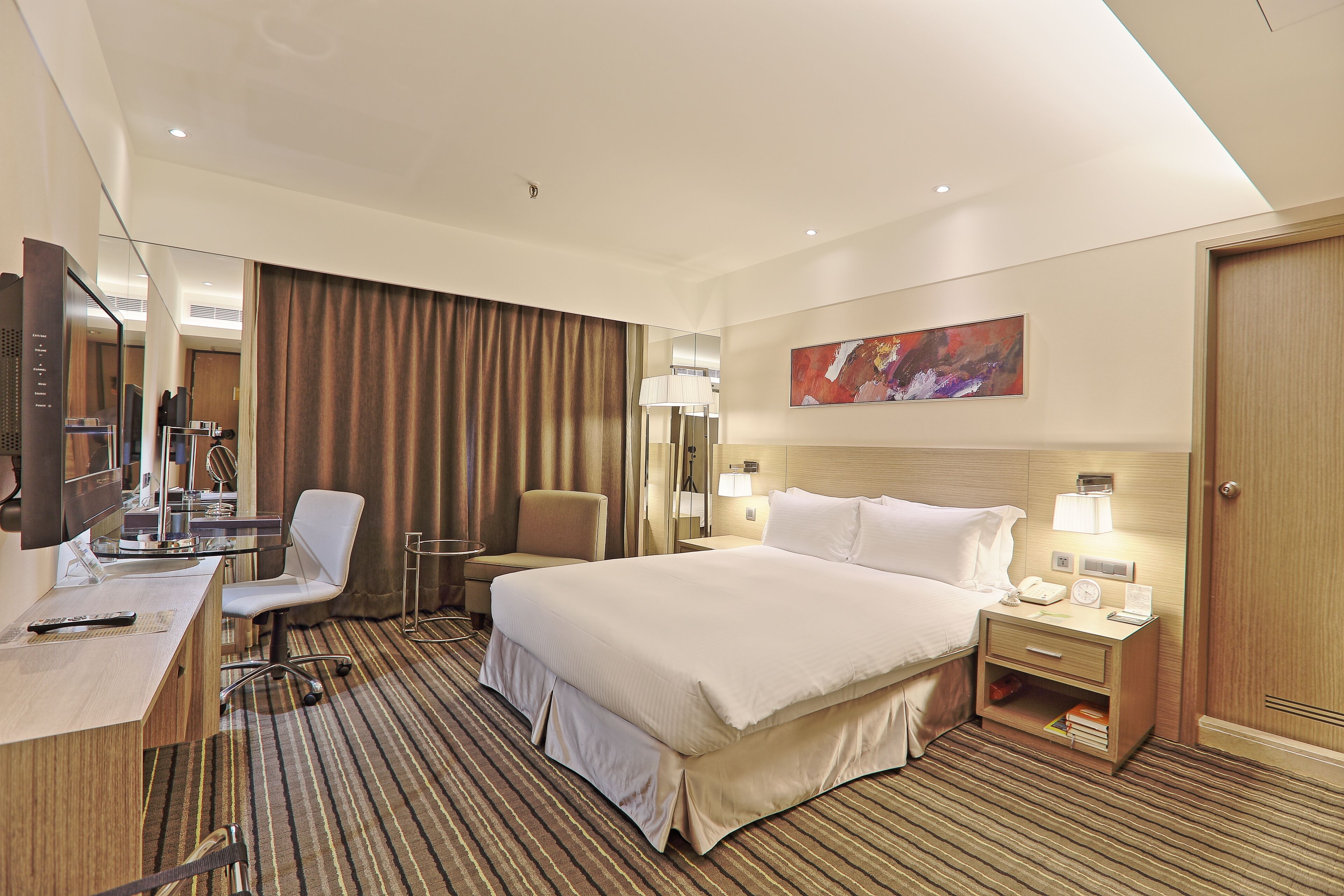 A hotel room with a large bed, a desk with a computer, a chair, a bedside table with a lamp, and a painting on the wall.