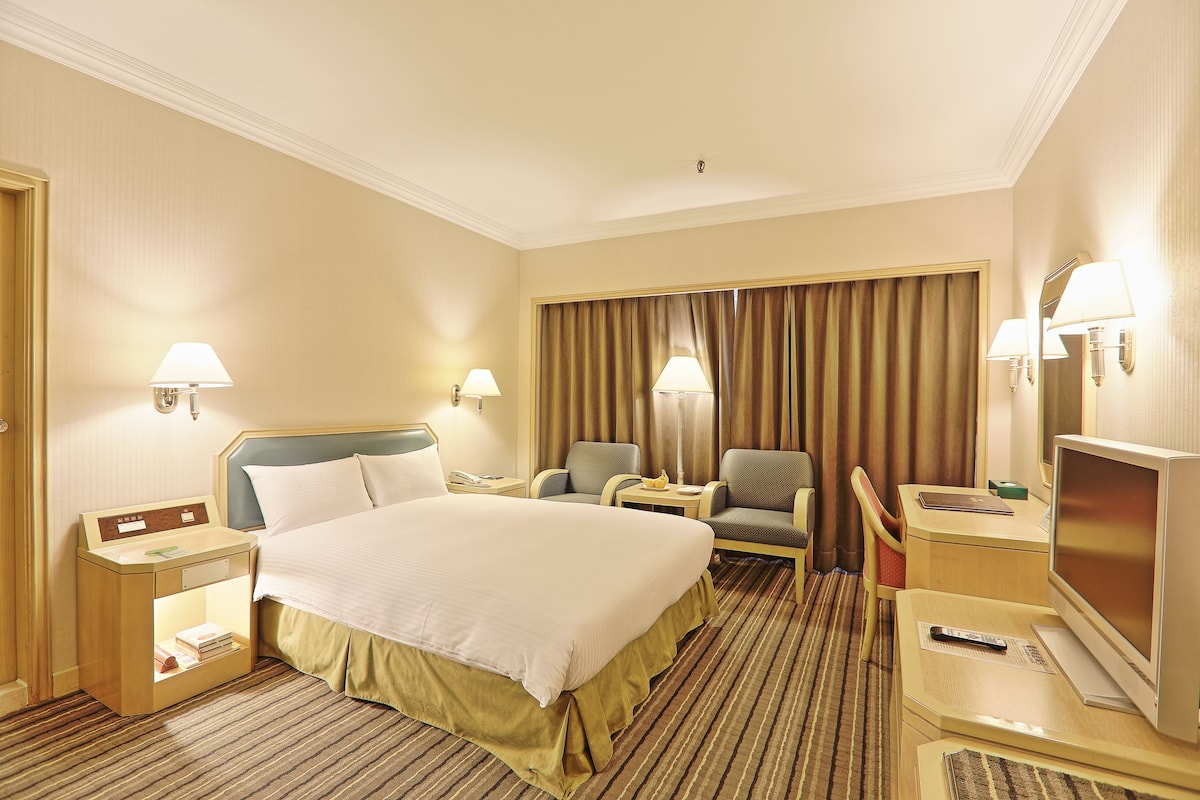 A hotel room with a large bed, two armchairs, a desk with a TV, and a bedside table with a lamp.