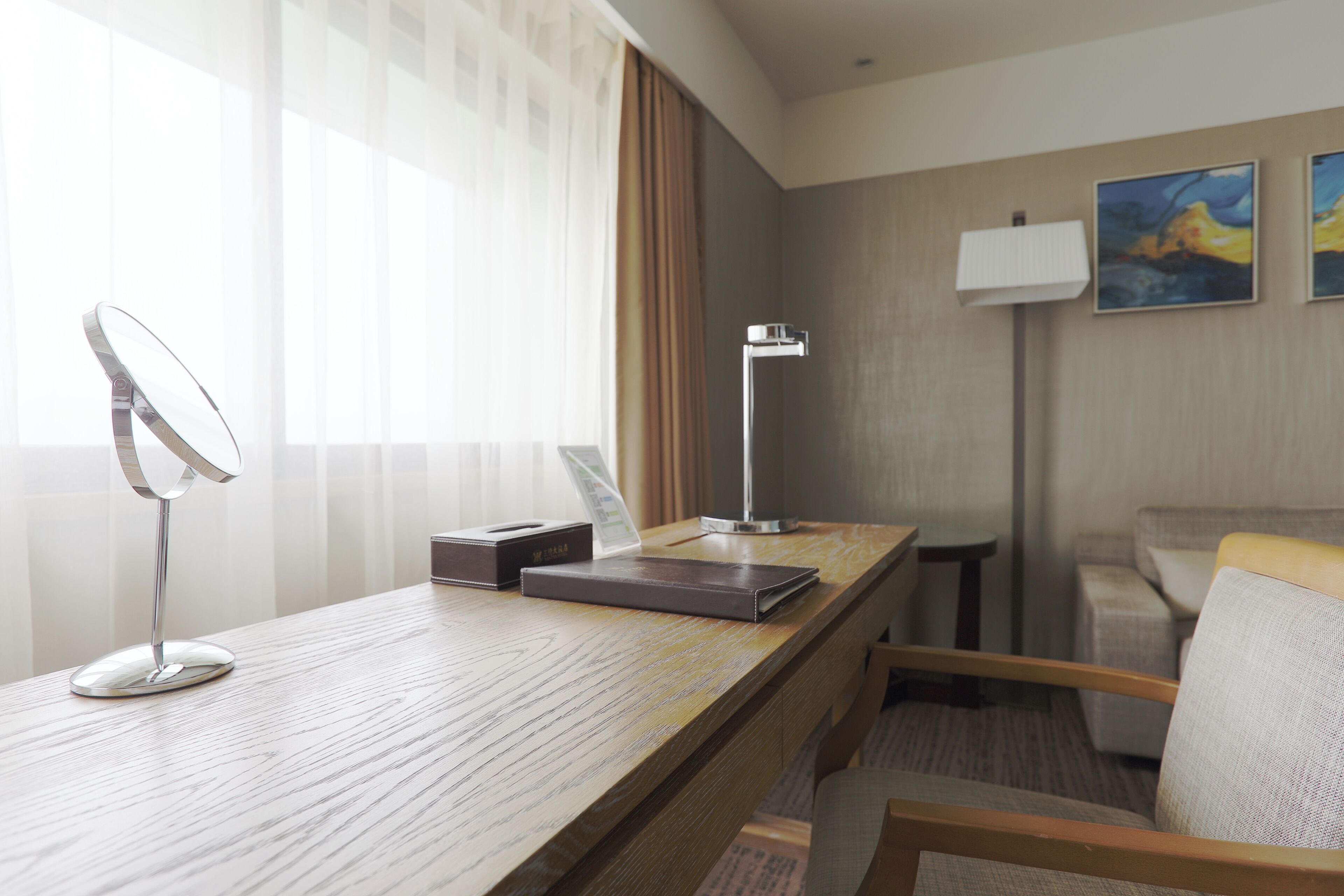 Habitación de hotel con escritorio de madera, lámpara moderna, una silla, un sofá y una obra de arte en la pared.