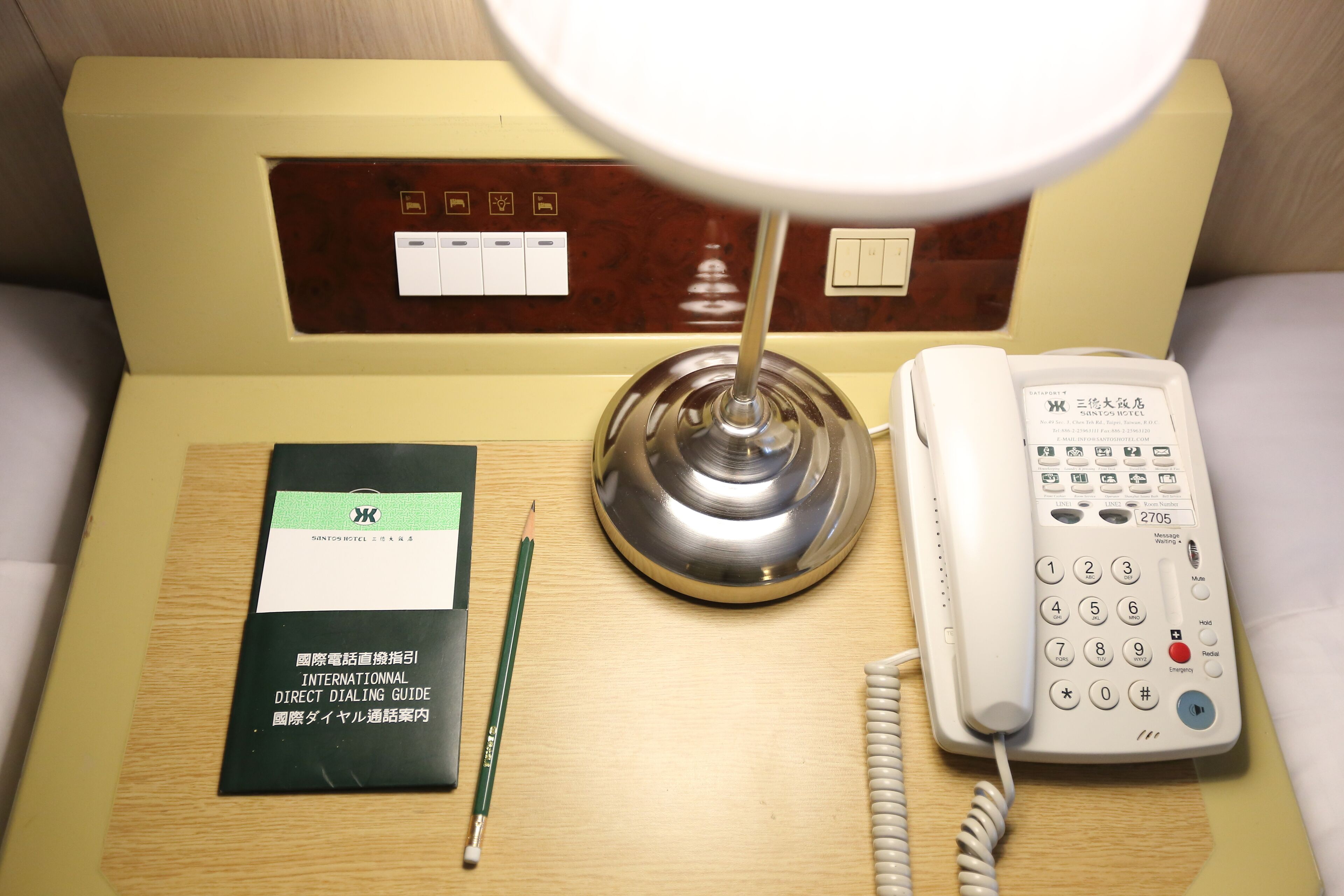 Een hotelkamer met nachtkastje, telefoon, lamp en een handleiding voor rechtstreeks bellen.