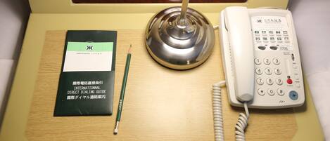 Een hotelkamer met nachtkastje, telefoon, lamp en een handleiding voor rechtstreeks bellen.