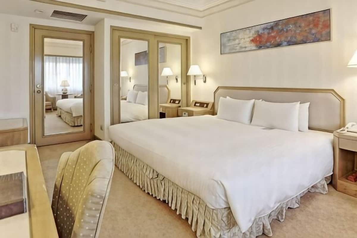 A hotel room with a large bed, a chair, a desk, a mirror, and a painting on the wall.