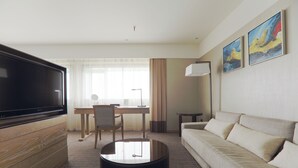 Quarto de hotel com televisão de ecrã plano, sofá, mesa de centro, secretária com cadeira e duas pinturas na parede.
