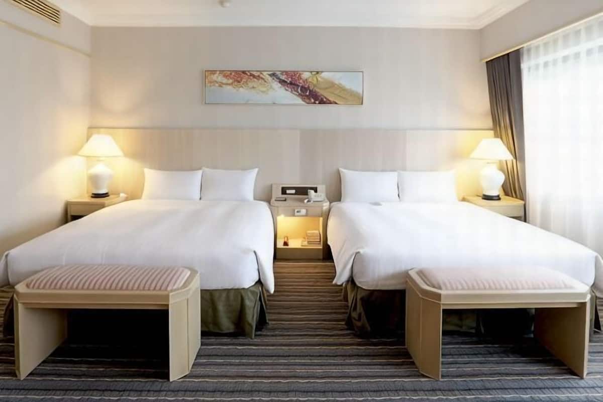 A hotel room with two beds, bedside tables with lamps, a small table between the beds, and a painting on the wall.