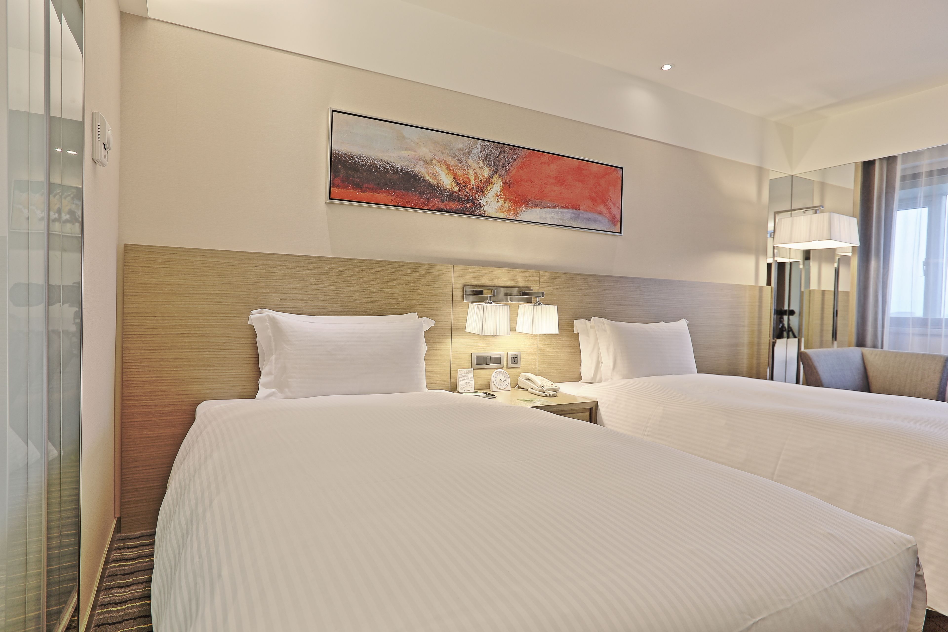 A hotel room with two beds, a large abstract painting, a bedside table with a lamp, and a view of the outside.
