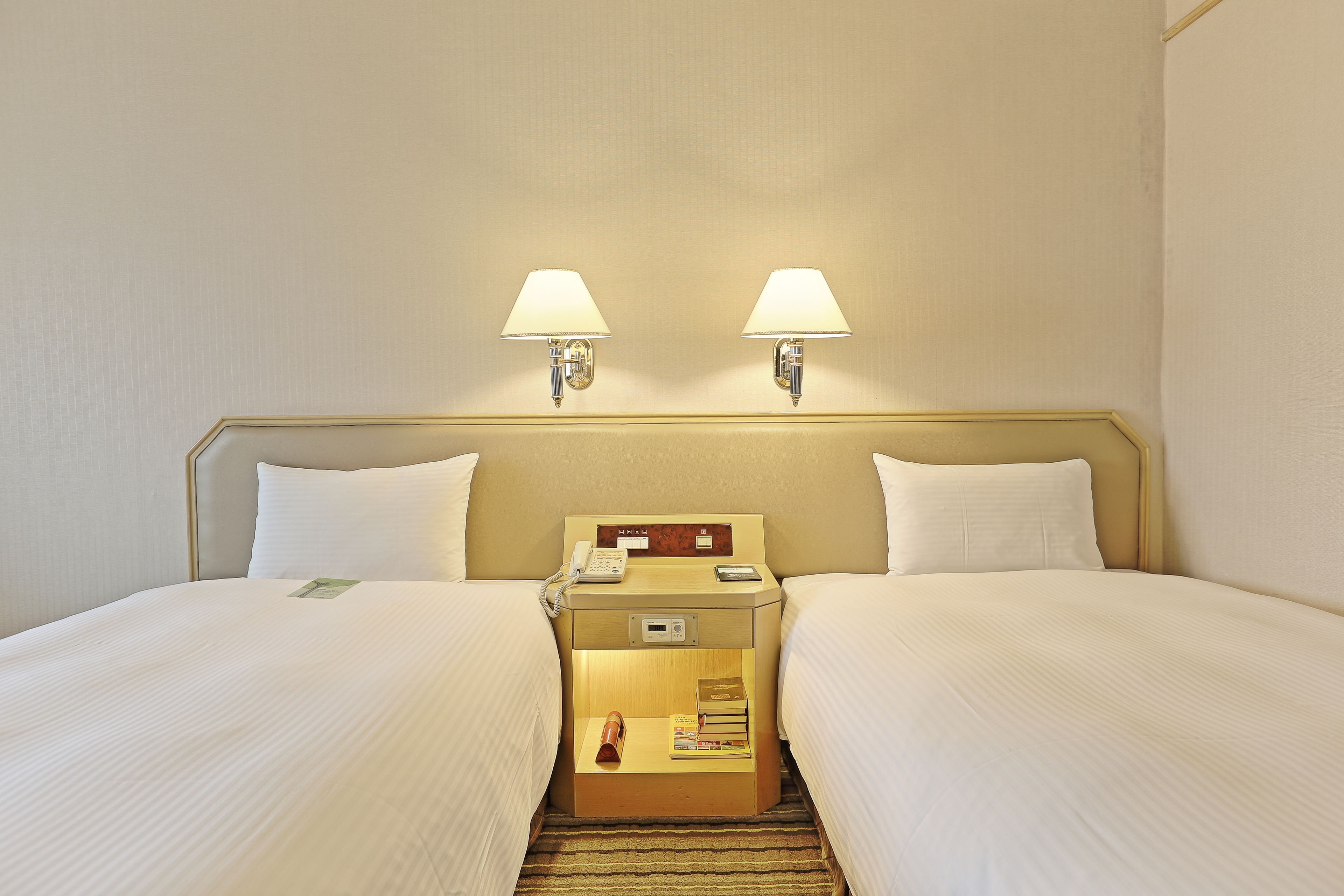 A hotel room with two beds, a bedside table with a lamp, a phone, and a small table with a book and a bottle.