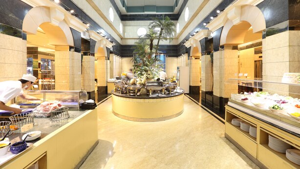Uma área de buffet com ilha central, rodeada por paredes de mármore e teto alto com iluminação embutida.