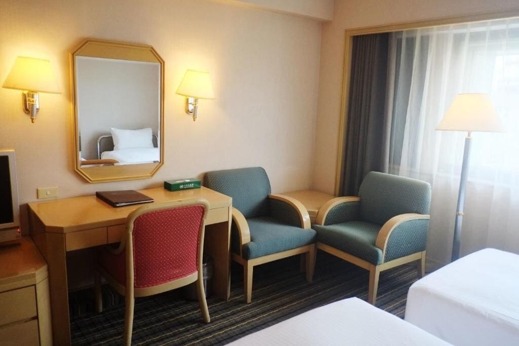 A hotel room with a desk, two chairs, a bed, a mirror, and two wall lamps.