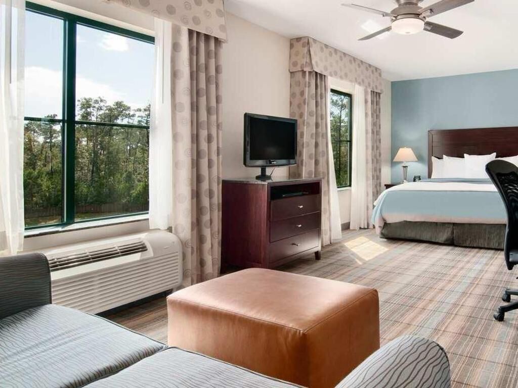 A hotel room with a bed, a television on a dresser, a sofa, an ottoman, and large windows with curtains.