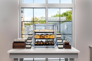 A bakery display case with various baked goods and a window view of a cityscape. - Homewood Suites Slidell (Slidell)