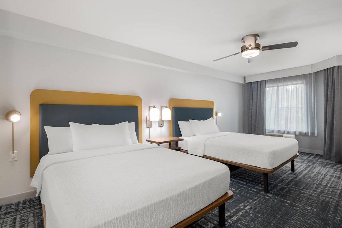 A hotel room with two beds, a ceiling fan, bedside lamps, and a large window with curtains.