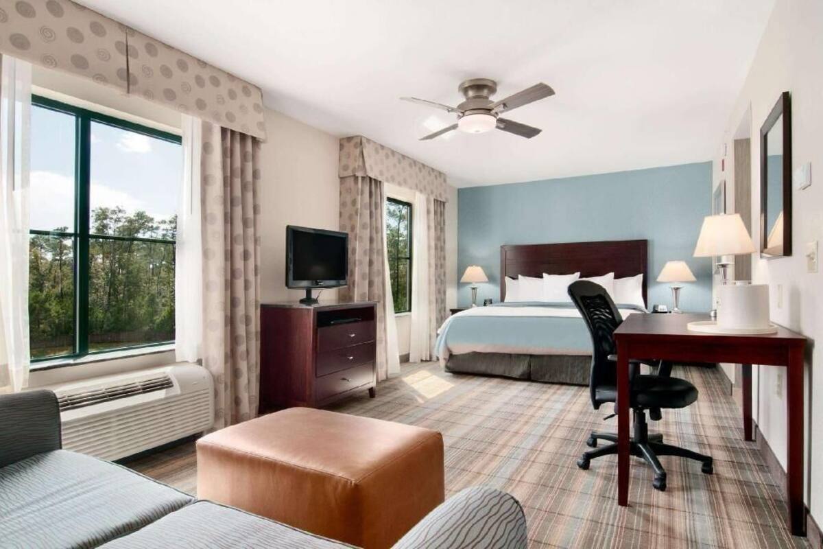 A hotel room with a bed, desk, chair, TV, and large windows.