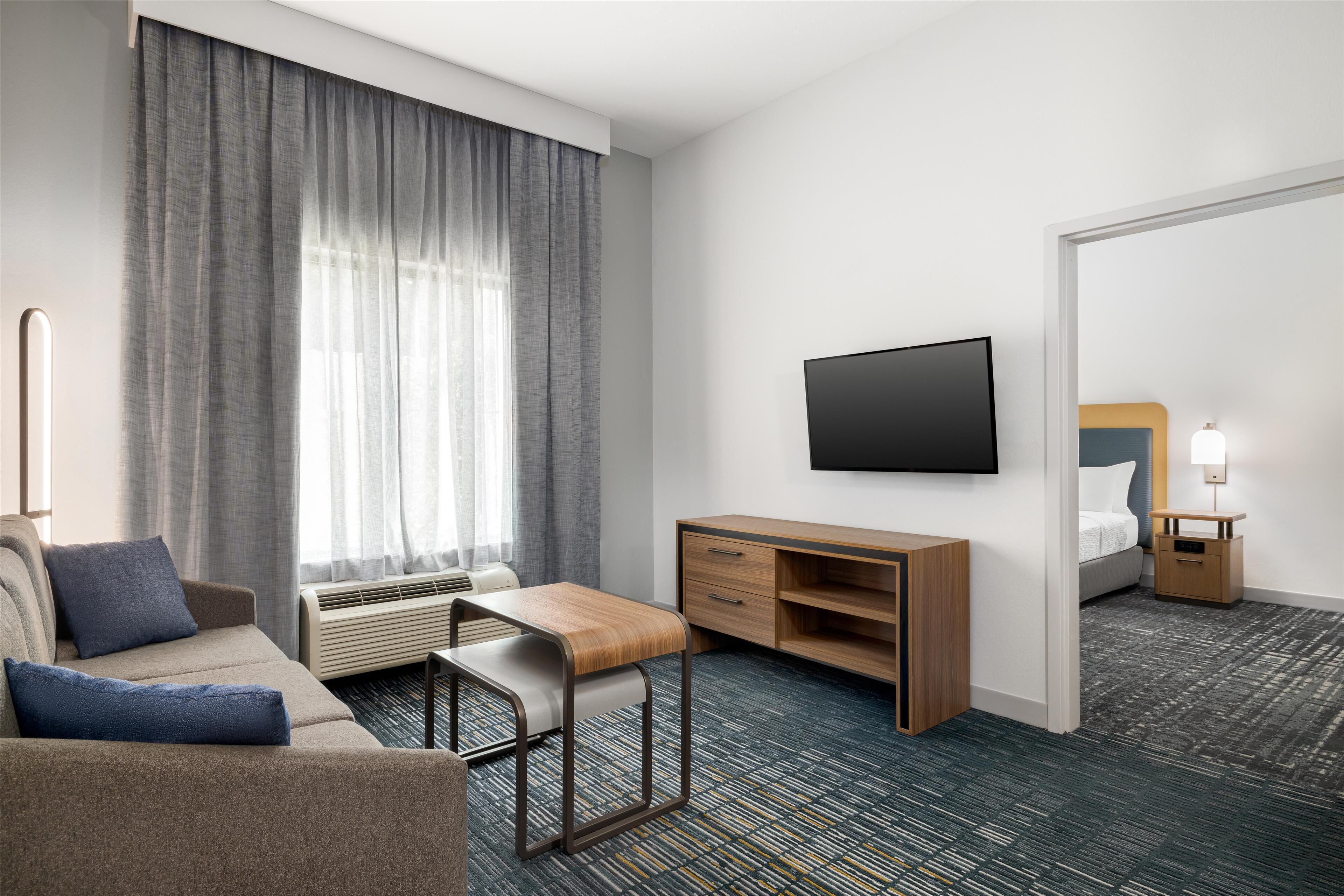 Ein Hotelzimmer mit einer Couch, einem Holztisch, einem Flachbildfernseher, einem Nachttisch mit Lampe und einem großen Spiegel.