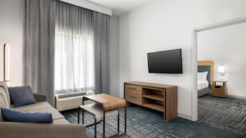 Habitación de hotel con sofá, un escritorio de madera, televisor de pantalla plana, mesita de noche con lámpara y un espejo grande.