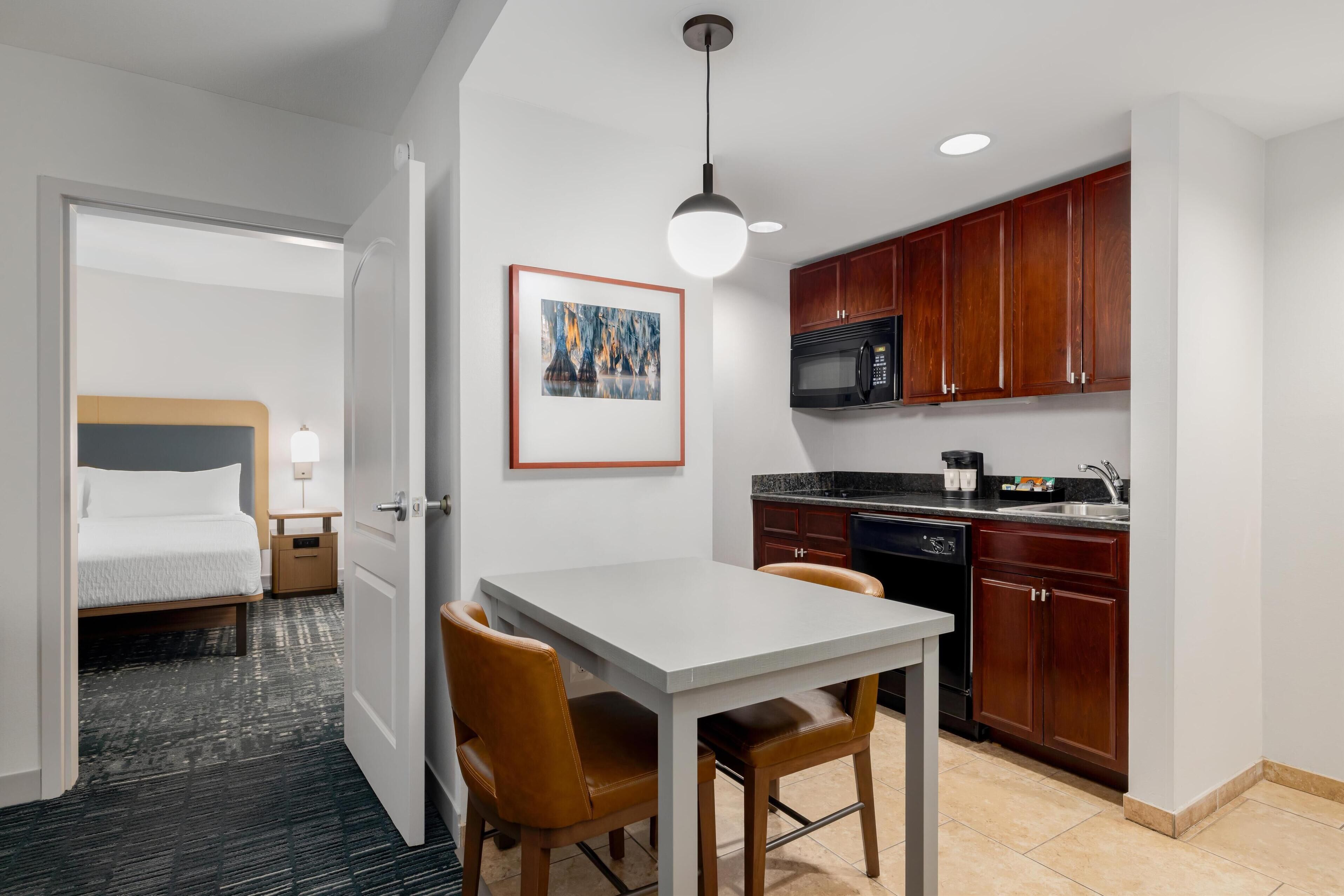 A compact hotel room with a bed, bedside table, lamp, and a view of the adjacent kitchen area.