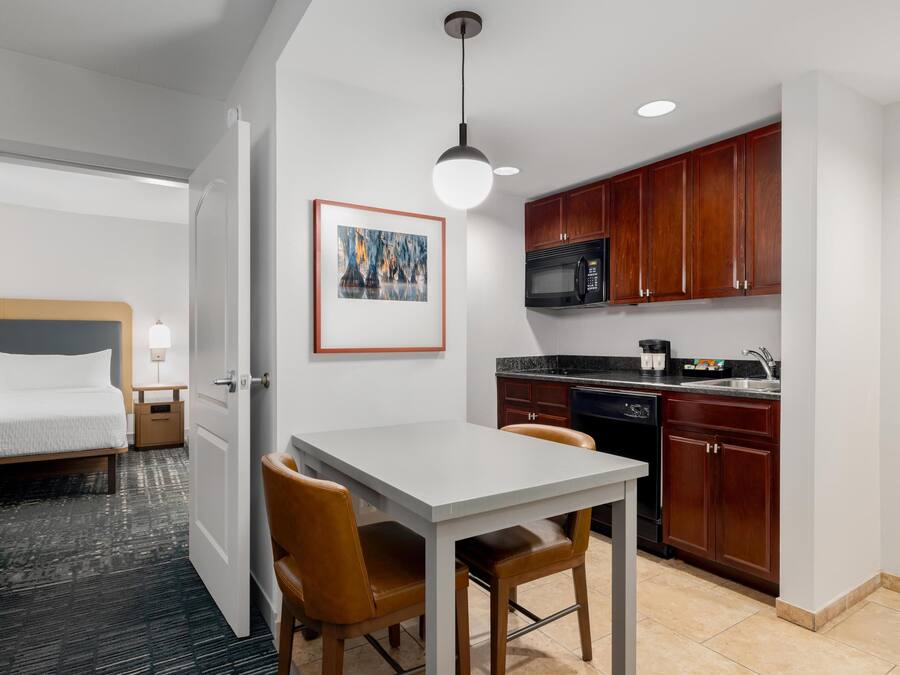 A compact hotel room with a bed, bedside table, lamp, and a view of the adjacent kitchen area.