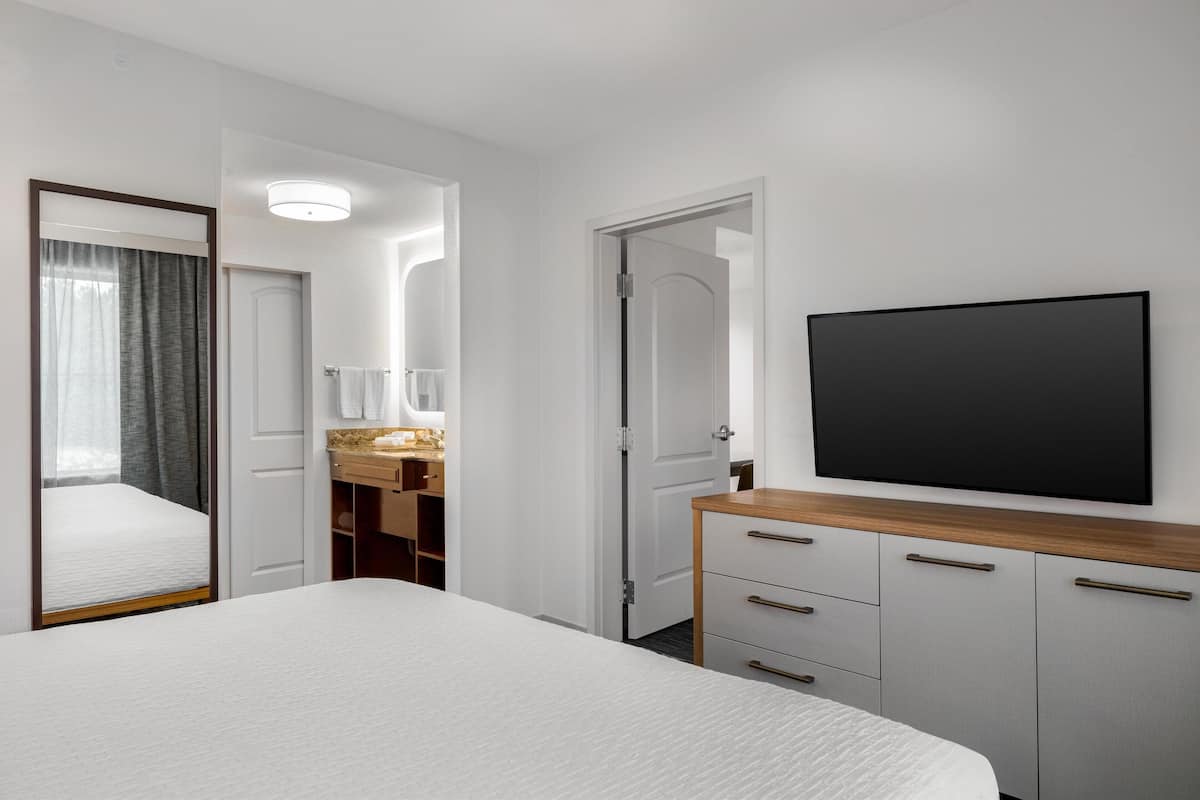 A hotel room with a bed, a TV mounted on the wall, a wooden dresser, and a bathroom visible through an open door.