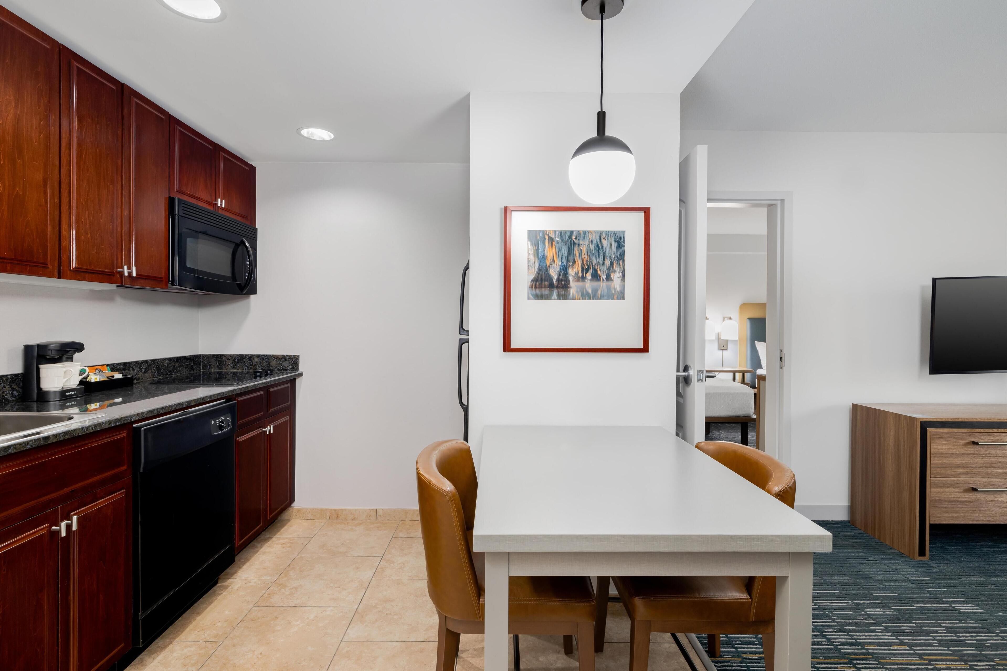 A modern kitchen with dark wood cabinets, a black microwave, and a dishwasher. There is a dining area with a white table and brown chairs. A framed artwork hangs on the wall, and a TV is mounted on the wall.