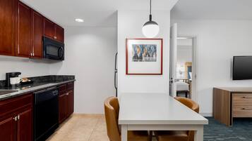 A modern kitchen with dark wood cabinets, a black microwave, and a dishwasher. There is a dining area with a white table and brown chairs. A framed artwork hangs on the wall, and a TV is mounted on the wall.