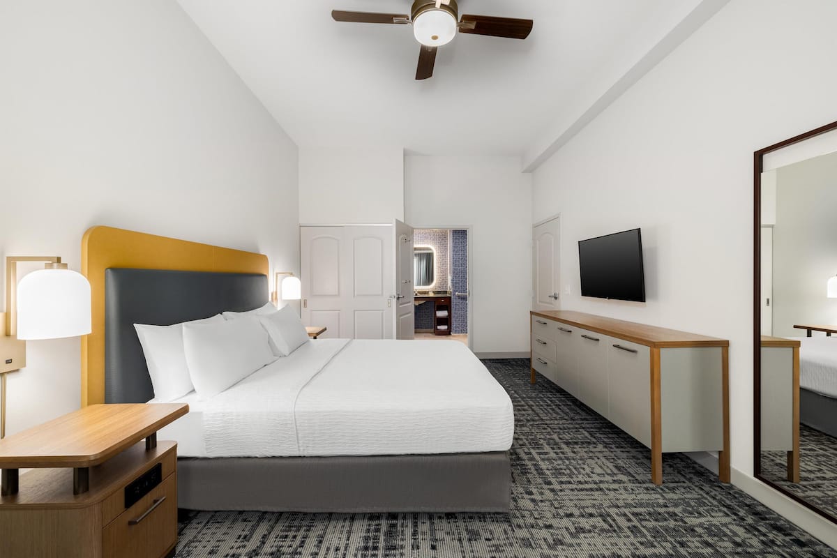 A hotel room with a bed, bedside table, lamp, TV, and a mirror.