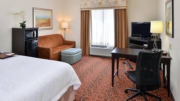 A hotel room with a bed, a sofa, a desk with a computer, a TV, and an air conditioning unit.