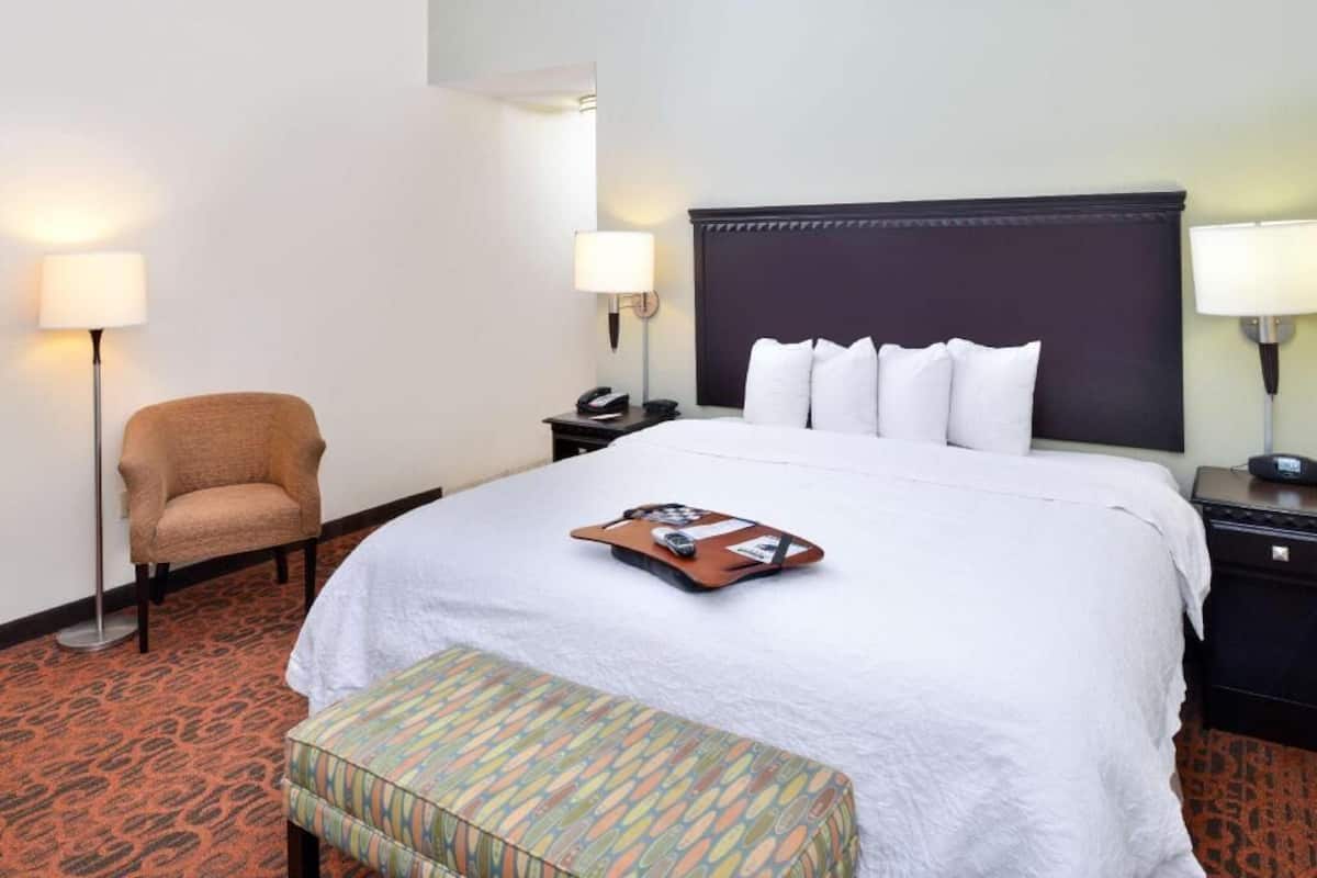 A hotel room with a bed, bedside tables, a chair, and a patterned bench.