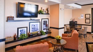 A hotel lobby with a television mounted on the wall, a seating area with chairs and a table, and framed pictures on the wall. - Hampton Inn Pecos (Pecos)