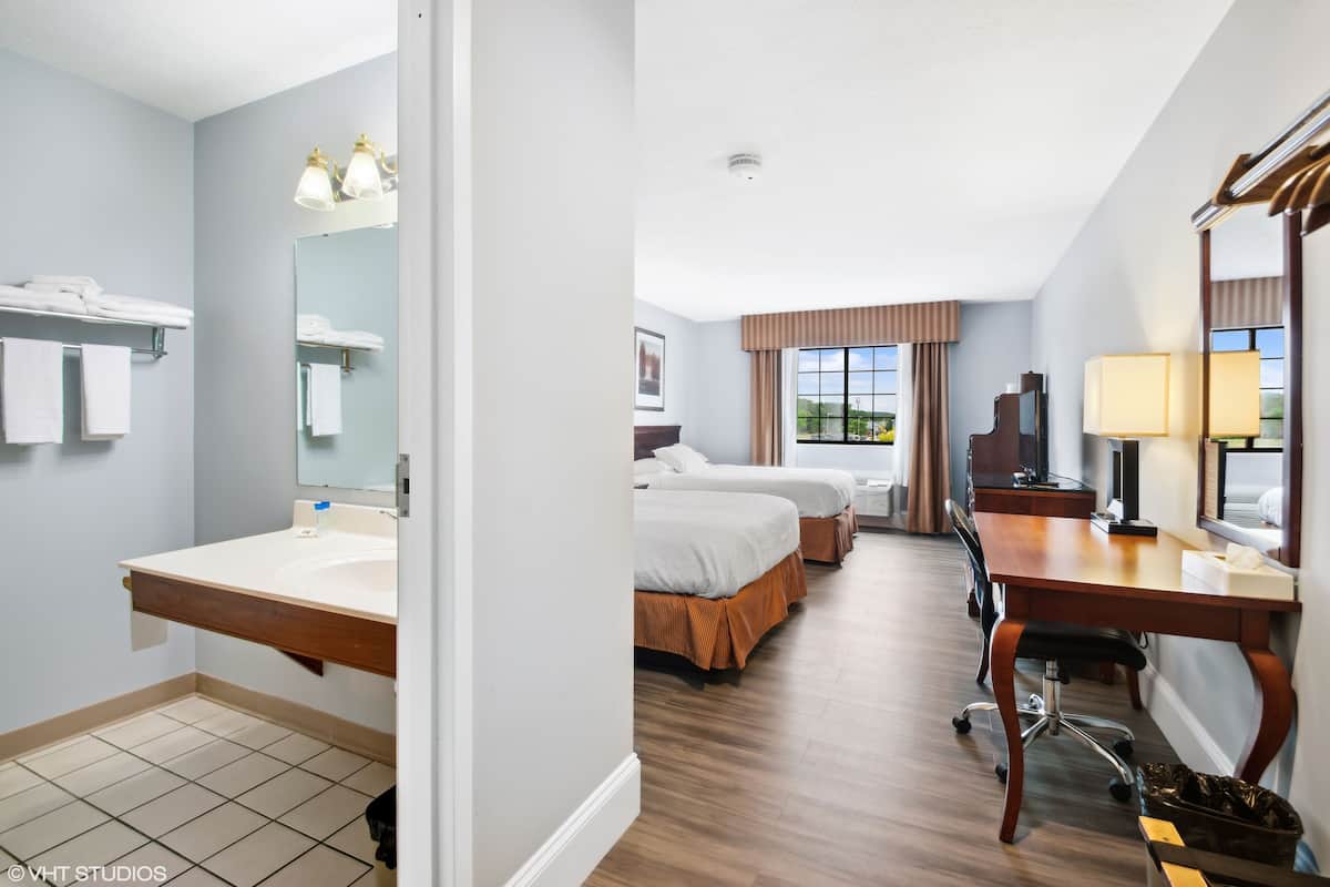 A hotel room with a large mirror, two beds, a desk with a chair, a lamp, and a window with curtains.