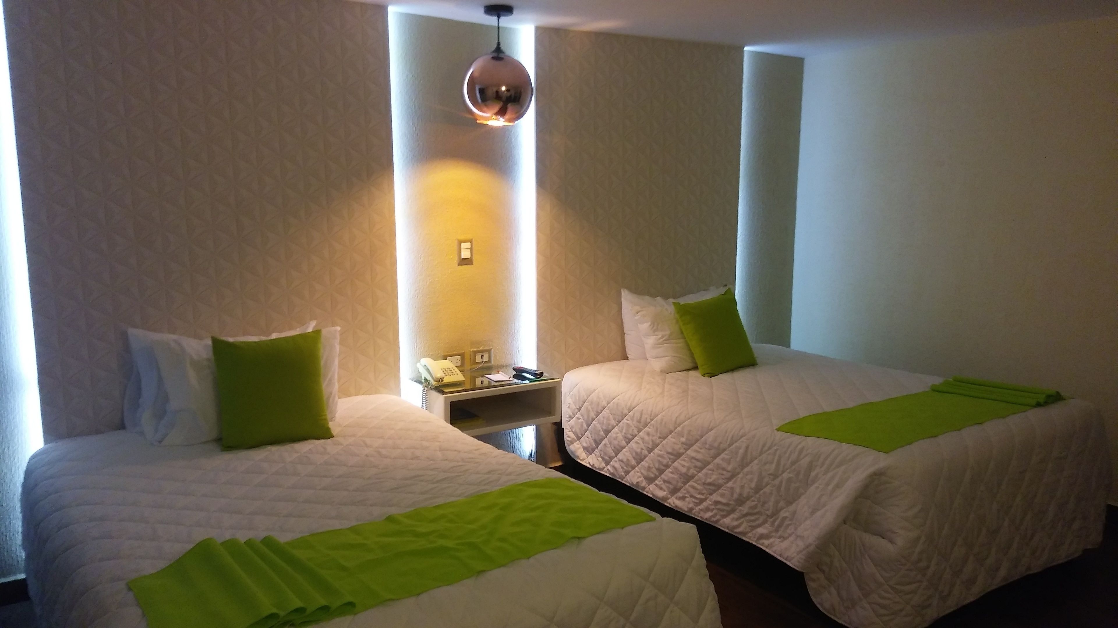 A hotel room with two beds, each with a green pillow and a white bedspread. There is a nightstand with a phone and a lamp between the beds.