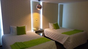 A hotel room with two beds, each with a green pillow and a white bedspread. There is a nightstand with a phone and a lamp between the beds.