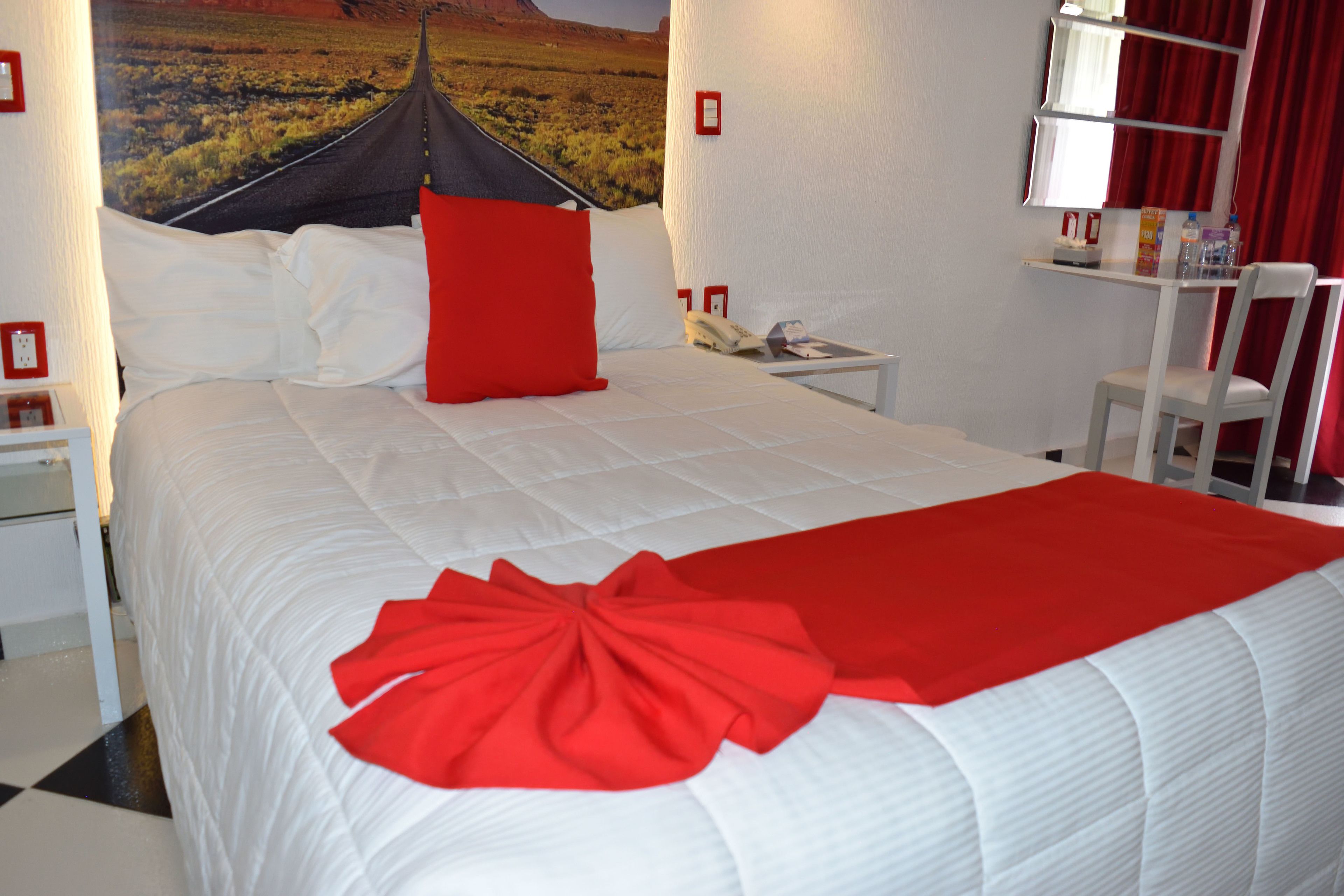 A neatly made bed with a red accent pillow and a red bow on the bedspread, a white nightstand with a telephone, and a red curtain on a window.