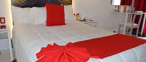 A neatly made bed with a red accent pillow and a red bow on the bedspread, a white nightstand with a telephone, and a red curtain on a window.